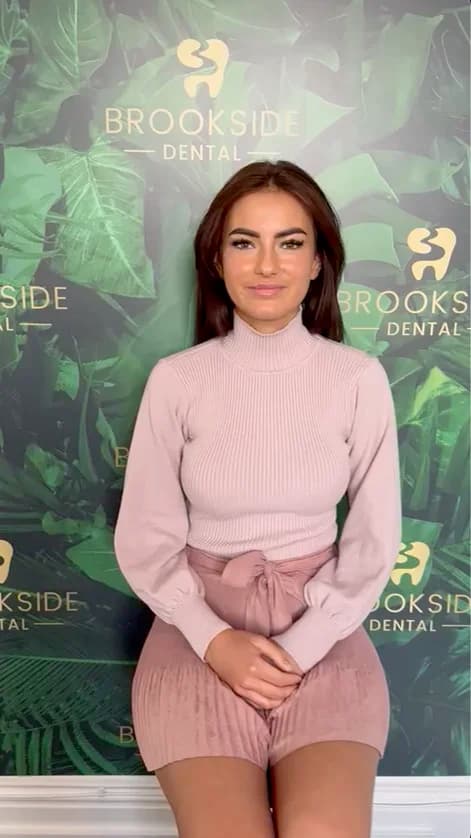 Woman with dark hair wearing a light pink turtleneck sweater and matching shorts sitting in front of a Brookside Dental backdrop with green leaves.