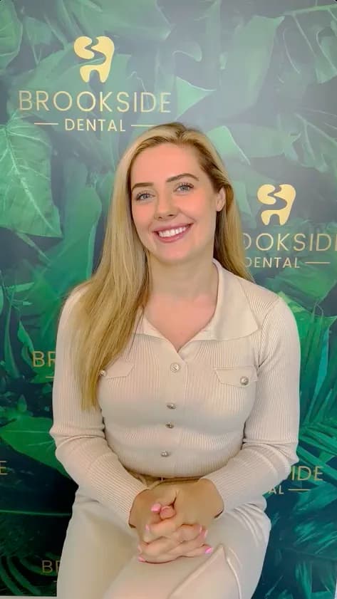 Smiling woman with long blonde hair sitting in front of a green leafy backdrop with Brookside Dental logos.