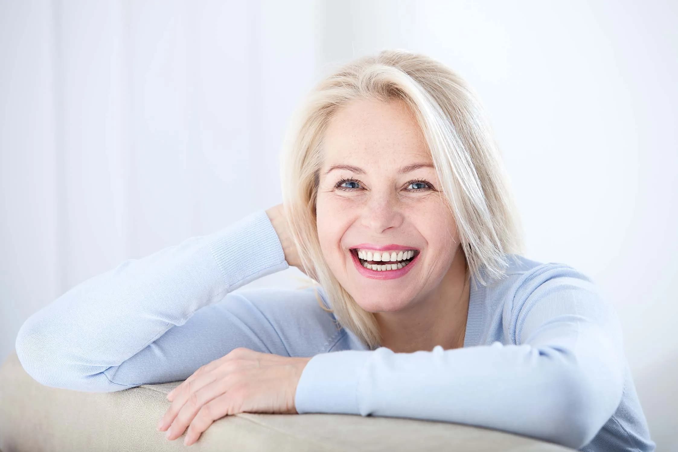 Smiling middle aged woman with blonde hair wearing a light blue sweater, leaning on a beige surface.