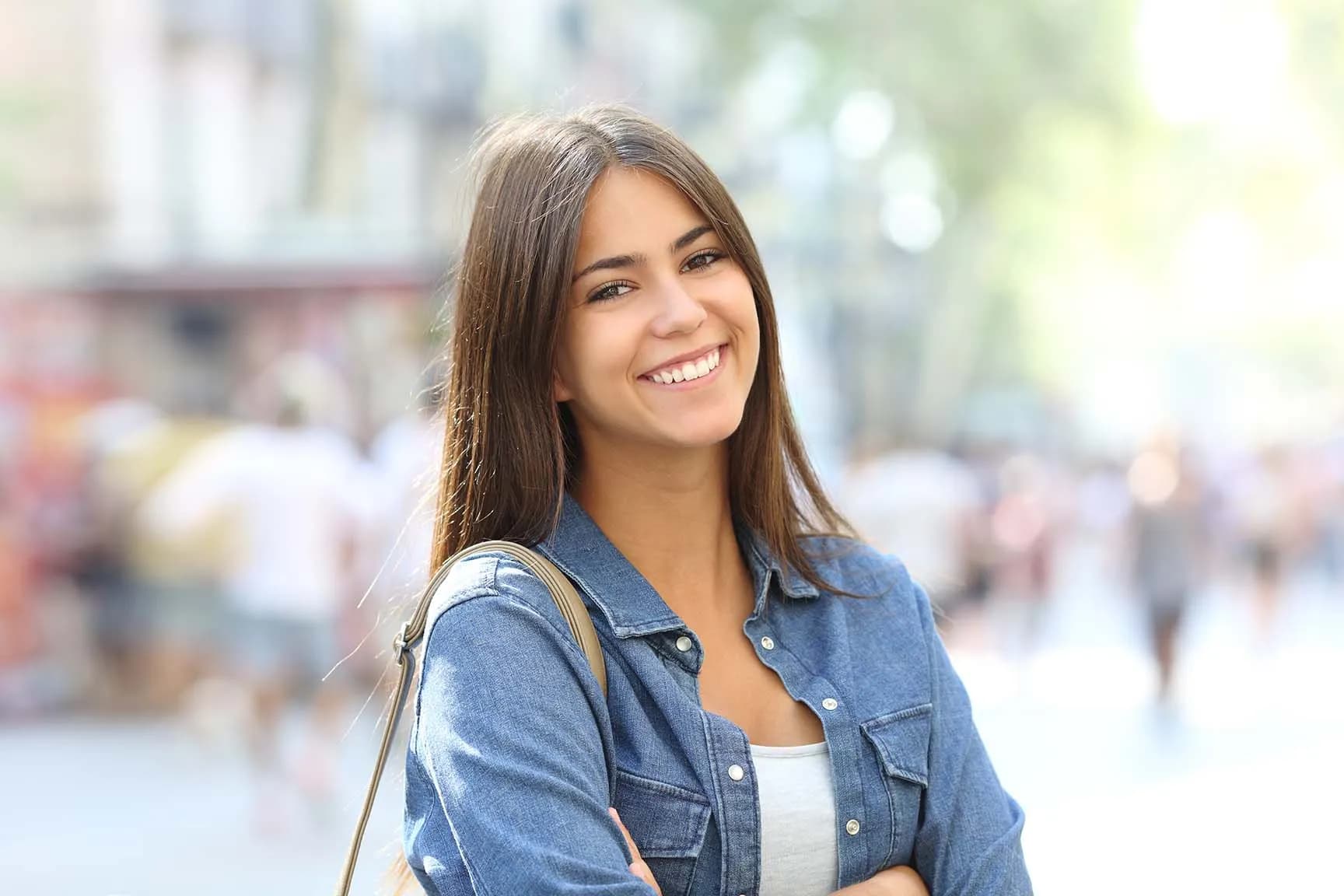 Smiling young woman with long brown hair wearing a denim jacket and white top outdoors.