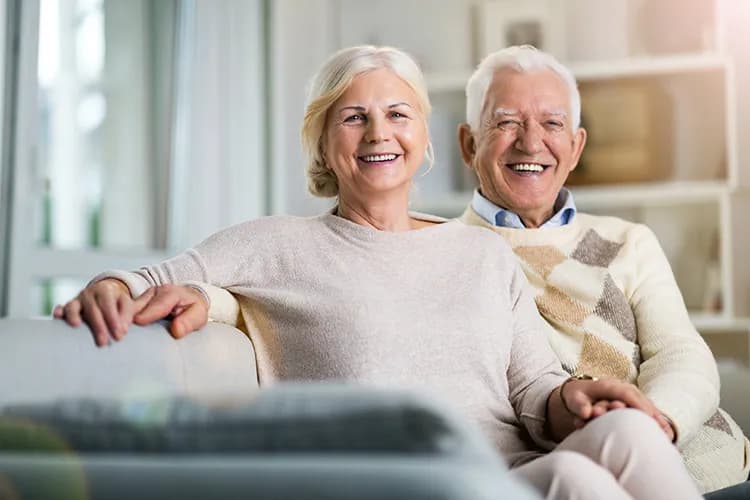 Smiling elderly couple sitting closely on a couch in a bright living room.