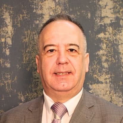 Middle aged man with short hair in a grey suit and striped tie against a textured dark wall.