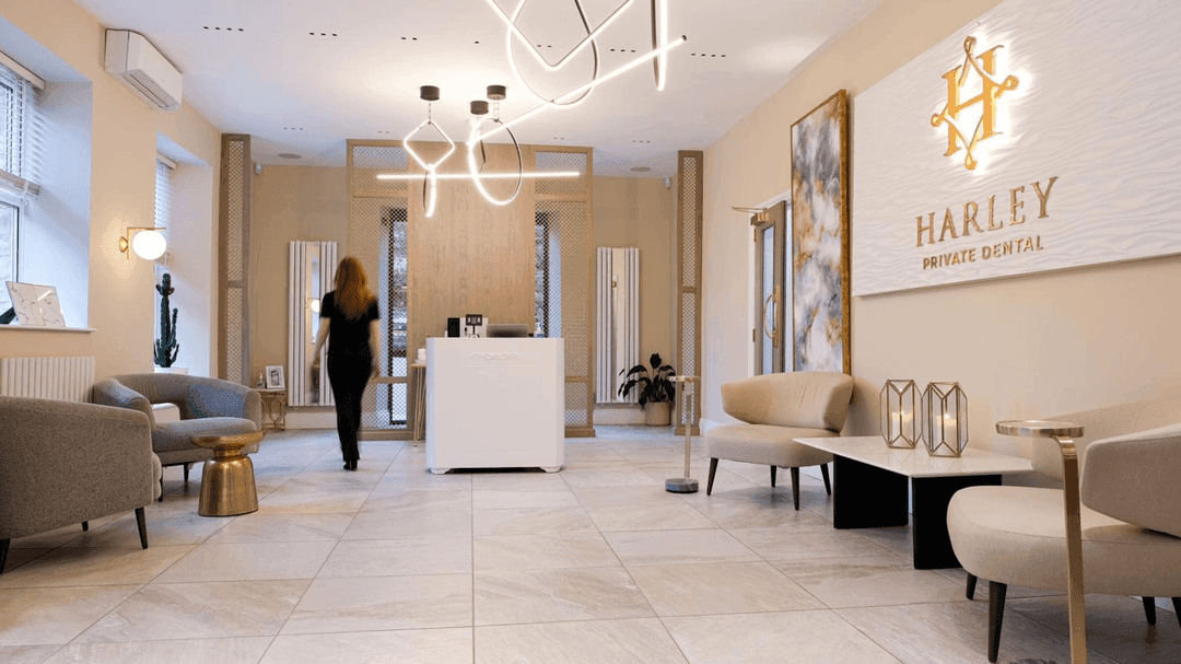 Modern dental clinic waiting area with beige walls, comfortable seating, contemporary lighting, and a reception desk with a person walking away.