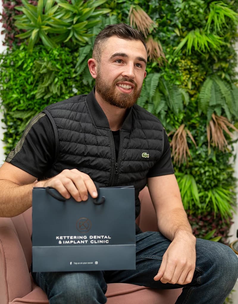 Smiling man sitting on a pink chair holding a Kettering Dental & Implant Clinic branded bag with a green leafy background.