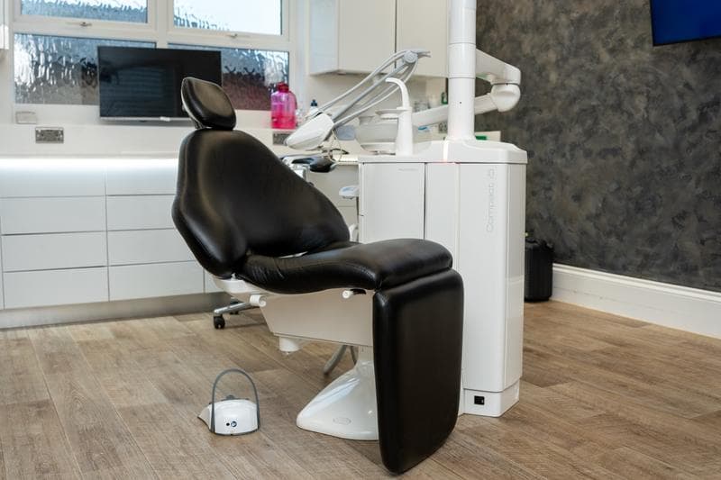 Black cushioned dental chair with attached dental equipment in a modern dental clinic room with wood flooring and white cabinets.