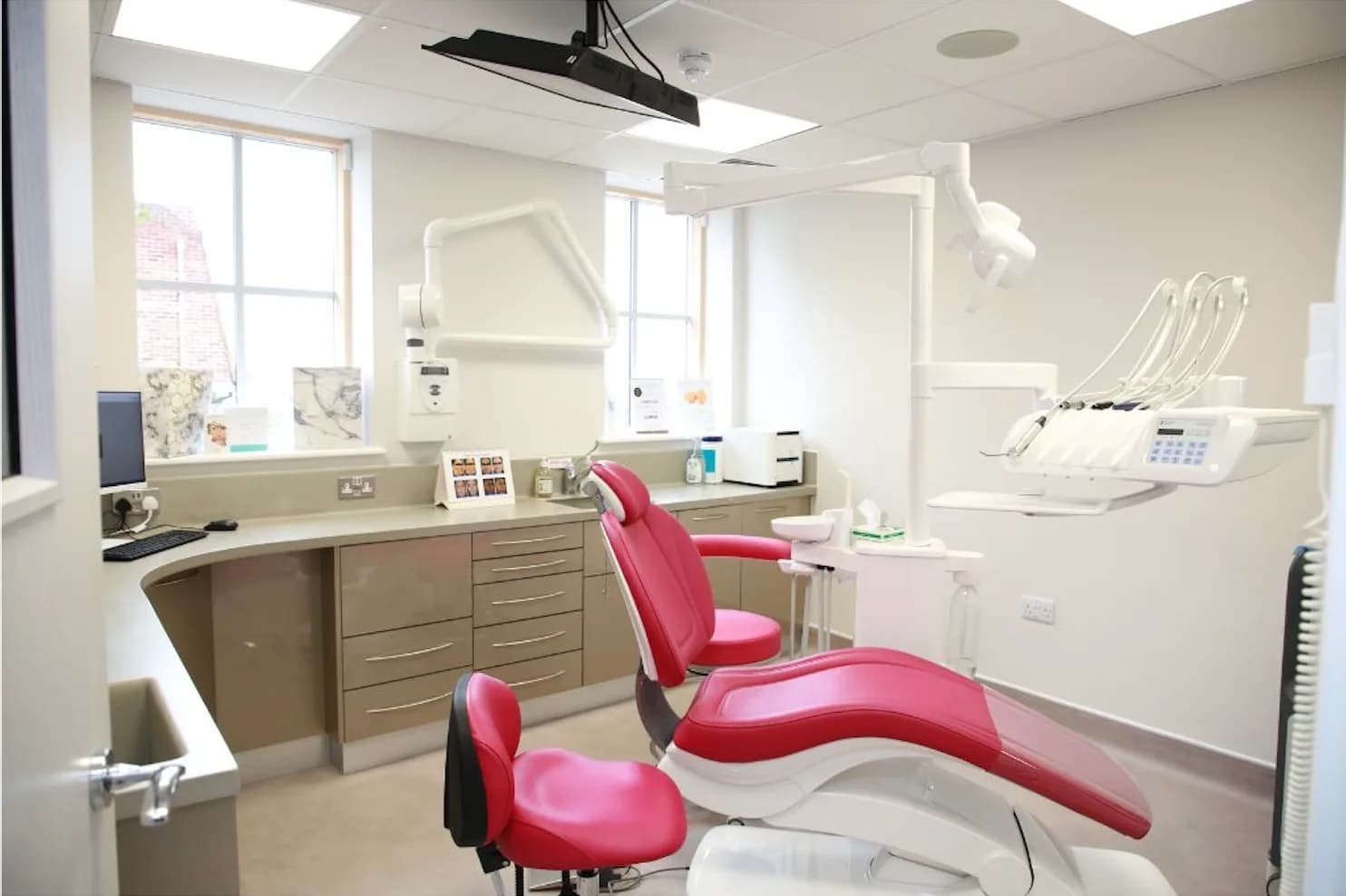 Bright dental treatment room featuring a red reclining dental chair, overhead examination lights, wall mounted instrument unit, cabinetry, and large windows providing natural light.