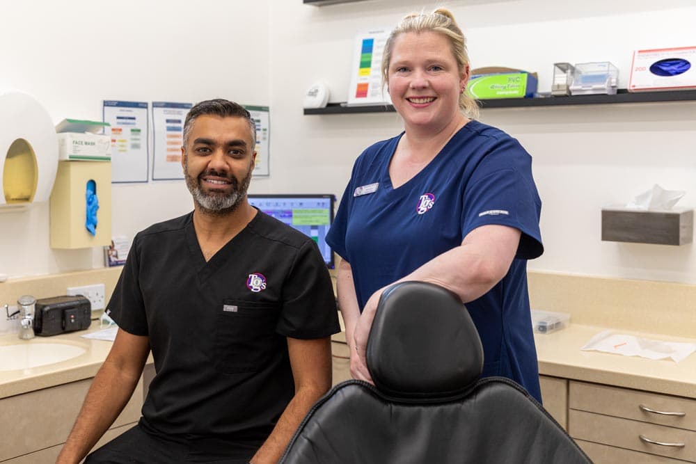 Two dental professionals smiling in a dental clinic room with dental equipment and supplies in the background.