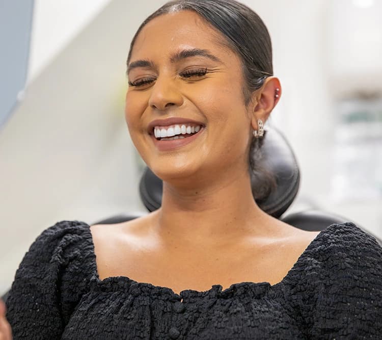 Smiling woman with eyes closed wearing a textured black top, sitting in a dental chair.
