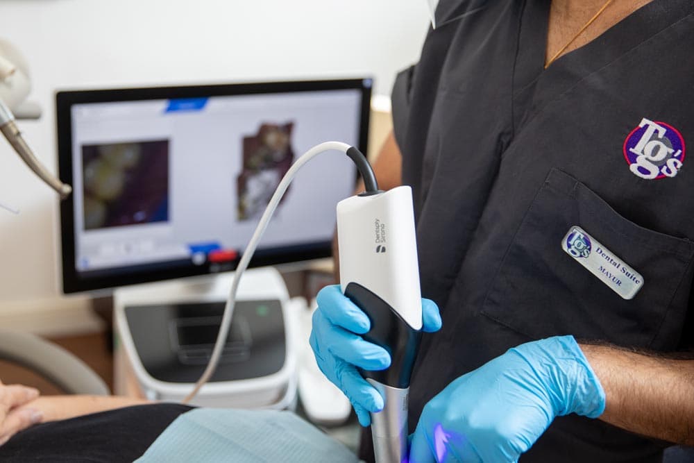 Dentist wearing blue gloves holding an intraoral digital scanner near a patient's mouth with a monitor displaying dental images in the background.